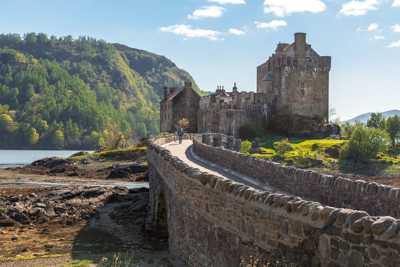 Long stone bridge leading up to Eilean Donan Castle with stone wall architecture and forested hills in the background