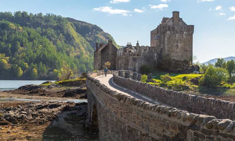 Long stone bridge leading up to Eilean Donan Castle with stone wall architecture and forested hills in the background