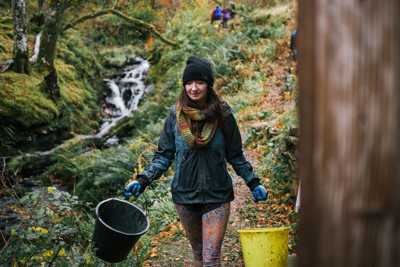 Young woman carrying two buckets through a lush, forested path in Tormore Forest