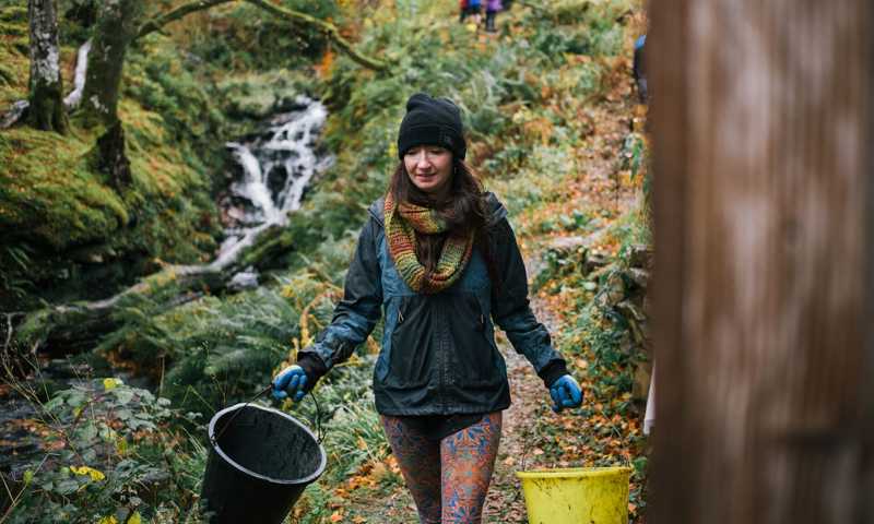 Young woman carrying two buckets through a lush, forested path in Tormore Forest
