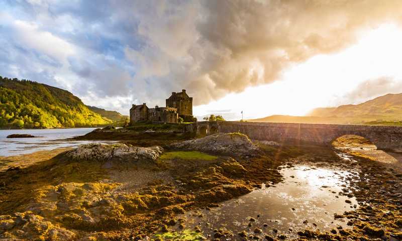 Eilean Donan Castle at sunset under dramatic sky with clouds and water puddles of the surrounding sea loch
