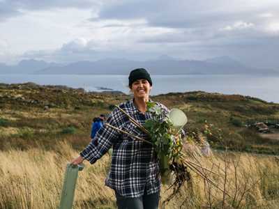 Young woman holding tree planting supplies on open ground in Tormore Forest with small islands in the background