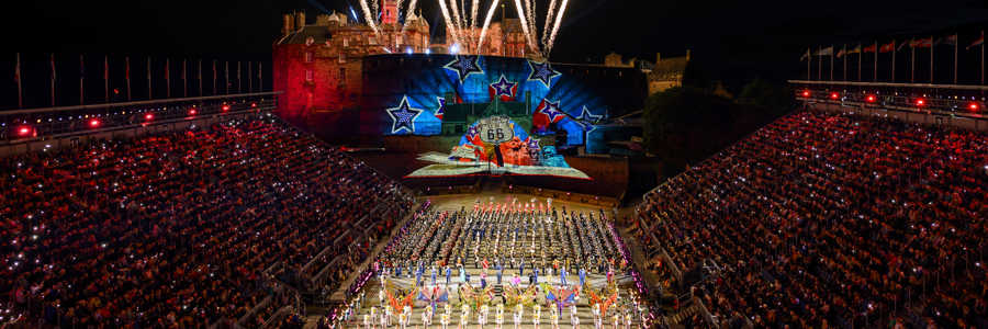 Performers in traditional military dress at the Royal Edinburgh Military Tattoo, set against the illuminated Edinburgh Castle at night with fireworks in the background