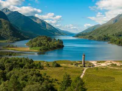 Large lake with clear blue water surrounded by lush green hills and a stone monument by the shores