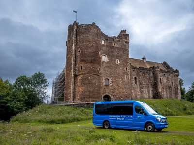 Blue Highland Explorer Mini Bus parked in front of Doune Castle with stone tower set against dramatic sky