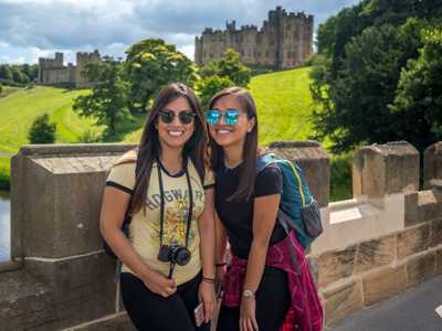 Two young women posing for a photo on a stone bridge leading towards Alnwick Castle