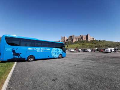 Blue Highland Explorer Tour bus parked before Bamburgh Castle on a sunny day