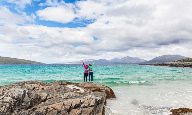 Two female travellers hugging and enjoying the view of turquoise waters by the shores of Isle of Harris