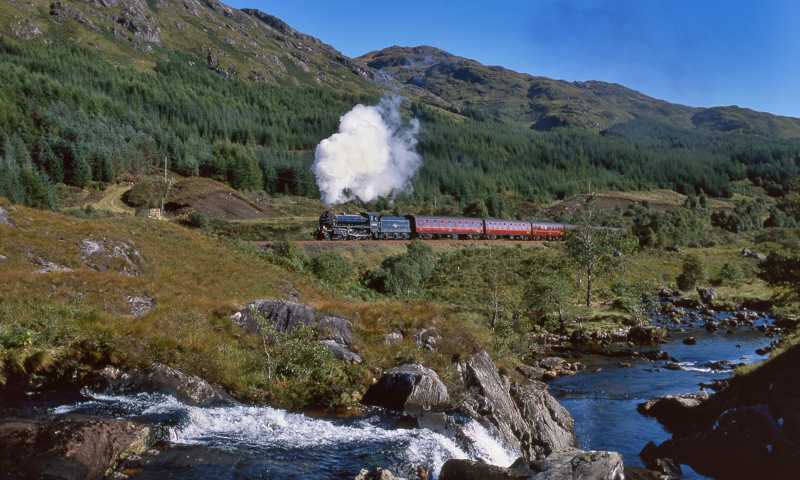 Jacobite Steam Train: red train carriages and black steam engine passing through forested area with rivers and hills